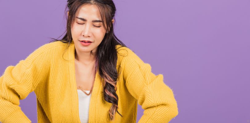 A woman in a yellow sweater standing against a purple background. Her hands are over her stomach as if in pain.